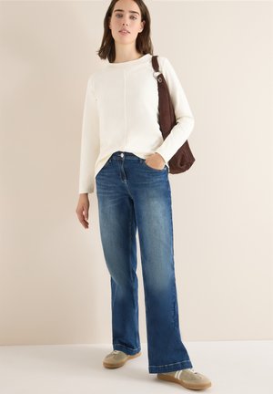 Haut beige à manches longues associé à un jean flare en denim bleu. L'ensemble comprend un sac à main marron et des baskets gris clair.