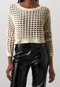 Kremowy sweter z luźnym splotem, szerokim dekoltem i długimi rękawami, w zestawieniu z błyszczącymi czarnymi spodniami w krokodyli wzór.