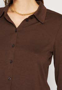 Femme portant une chemise marron boutonnée avec un col et un collier chaîne dorée autour du cou.