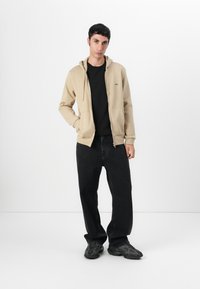Felpa beige con zip, tasche a marsupio e cappuccio con cordino, indossata sopra una maglietta nera, abbinata a pantaloni neri larghi e sneaker scure.