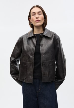 Frau trägt dunkelbraune, lederne Jacke mit Knöpfen über einem schwarzen Top und blauen Jeans, steht mit den Händen hinter dem Rücken vor einem einfarbigen Hintergrund.