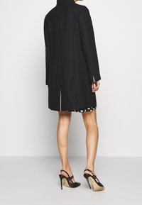 Black coat with a high collar and long sleeves, featuring a single back vent and contrasting seam details, paired with a patterned dress.