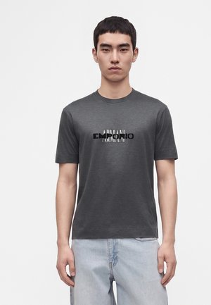 Jeune homme aux cheveux courts noirs portant un t-shirt Armani gris foncé et un jean bleu clair, debout devant un fond blanc uni.