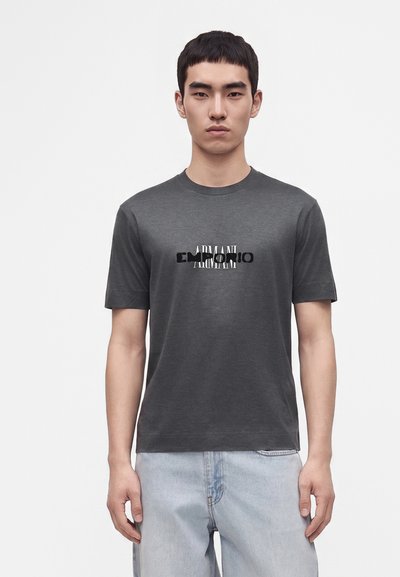 Млад мъж с къса черна коса, носещ тъмносива тениска Armani и светлосини дънки, стоящ на бял фон.