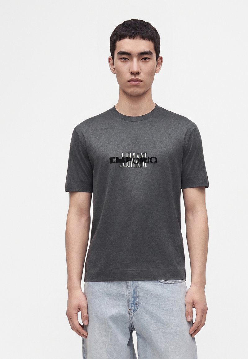 Jeune homme aux cheveux courts noirs portant un t-shirt Armani gris foncé et un jean bleu clair, debout devant un fond blanc uni.