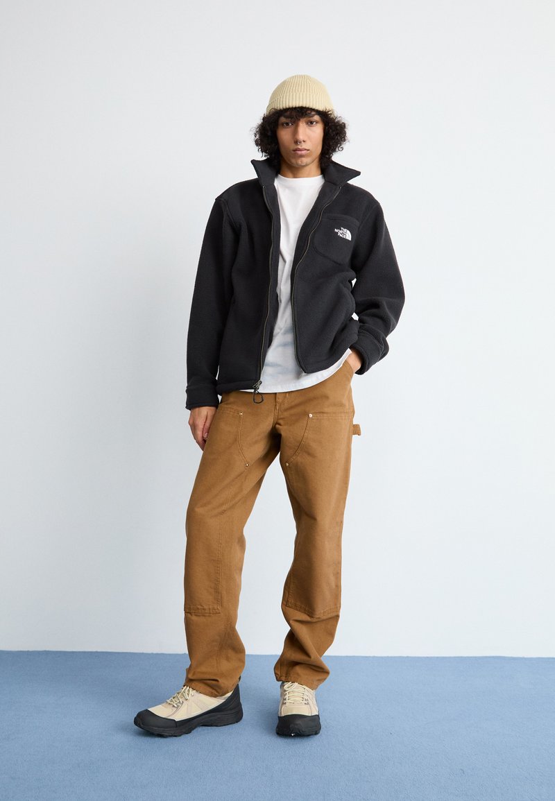 Veste en polaire noire avec zip, bonnet beige, t-shirt blanc, pantalon en toile marron et chaussures de randonnée beige/noir. Design simple et coupe décontractée.