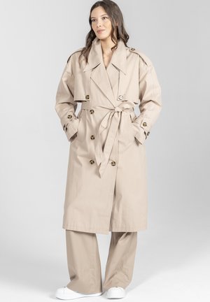 Kobieta w beżowym płaszczu trencz z paskiem związanym w talii, do tego spodnie o szerokich nogawkach w tym samym kolorze oraz białe trampki, stojąca na tle o neutralnym kolorze.