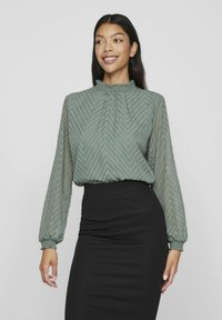 Kobieta w półprzezroczystej, długiej bluzce z długim rękawem w zielonym kolorze, z wzorem w zygzaki i falbaniastym kołnierzykiem, w zestawieniu z czarną spódnicą o wysokim stan.