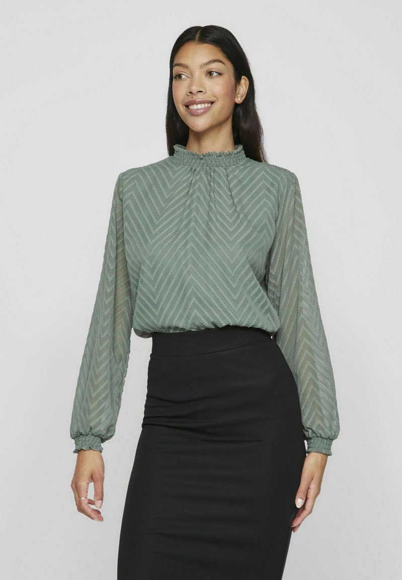 Kobieta w półprzezroczystej, długiej bluzce z długim rękawem w zielonym kolorze, z wzorem w zygzaki i falbaniastym kołnierzykiem, w zestawieniu z czarną spódnicą o wysokim stan.