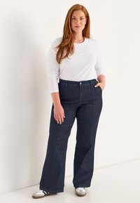 Nainen, jolla on pitkät punaiset hiukset, pukeutuneena valkoiseen pitkähihaiseen paita, tummansiniset pöksyt ja valkoiset tennarit, seisoo tasaisella seinällä.