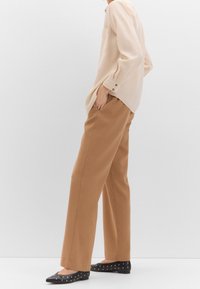 Chemise beige à manches longues avec une poche, associée à un pantalon camel à jambes larges. Des ballerines noires ornées de clous dorés complètent la tenue.
