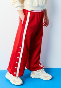 Røde sweatpants med hvide sidelister, med trykknapper langs den ydre søm. Kombineret med beige sneakers, sat mod et blåt gulv.