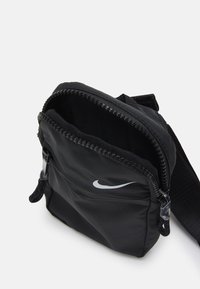 Černá nylonová crossbody taška se zipovým uzávěrem, s výrazným stříbrným logem Nike a nastavitelným popruhem pro všestrannost.