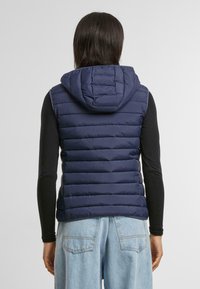 Gilet matelassé bleu marine avec une capuche, présentant un matelassage horizontal et une silhouette ajustée. Porté par-dessus un haut noir à manches longues.