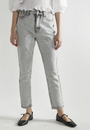 Donna che indossa jeans a vita alta grigio chiaro effetto acid wash, top bianco con maniche a sbuffo e ballerine nere Mary Jane, in piedi davanti a uno sfondo semplice.