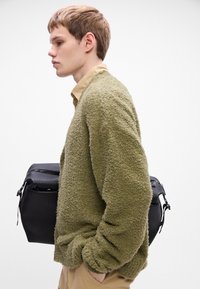 Groene gestructureerde trui met een losse pasvorm, gedragen over een beige shirt, gecombineerd met een zwarte tas met een ritsluiting en een gestructureerde vorm.