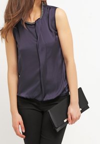 Navy silk sleeveless top with a subtle sheen, featuring a beaded neckline. Paired with a black clutch, held in both hands.