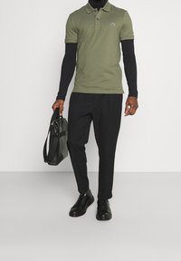 Polo vert olive avec deux boutons et logo Lacoste, associé à un pantalon noir fuselé et des baskets noires texturées, tenant un sac gris.