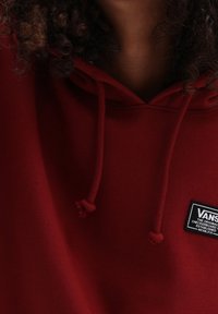Persona de cabello rizado que lleva una sudadera roja de Vans con cordones y un parche negro del logo de Vans en el pecho.