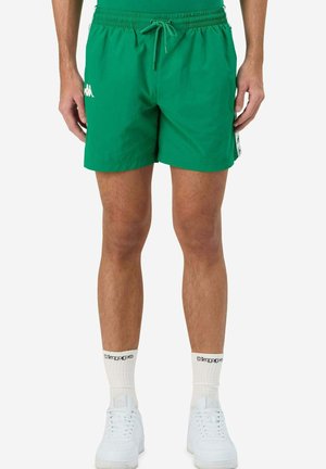 Pantaloni da nuoto verdi con vita elastica, lacci e tasche laterali; presenta il logo bianco del marchio sul lato sinistro. Indossati con sneakers bianche.