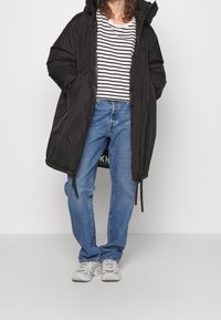 Černý péřový kabát s kapucí, v kombinaci s proužkovaným tričkem, modrými džínami a šedými teniskami. Kabát obsahuje detaily se šňůrkami.