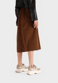 Gonna midi in velluto a coste marrone abbinata a sneakers beige. La giacca è in pelle nera con dettagli con zip, creando un look a strati.