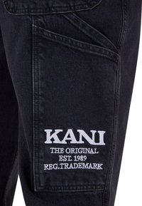 Svarte denimbukser med et stort, hvitt brodert logo som sier "KANI," med ekstra tekst nedenfor. Distressed lommepreg er synlig.