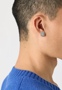 Orecchino quadrato d'argento con dettagli intricati, indossato nell'orecchio sinistro. Maglione blu con una scollatura a costine visibile.