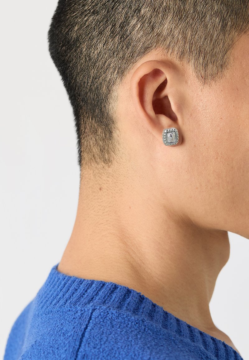 Orecchino quadrato d'argento con dettagli intricati, indossato nell'orecchio sinistro. Maglione blu con una scollatura a costine visibile.