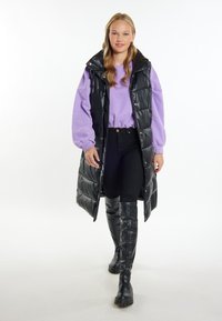 Schwarze, voluminöse ärmellose Jacke über einem lavendelfarbenen Sweatshirt, kombiniert mit schwarzen Skinny-Jeans und kniehohen schwarzen Stiefeln mit glänzender Oberfläche.