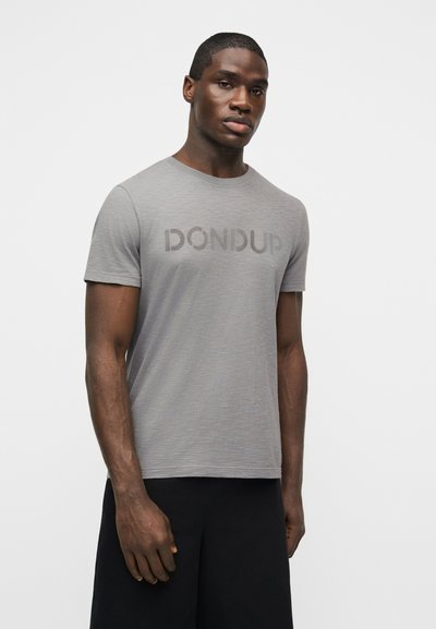 Άνδρας με γκρι φανέλα Dondup και μαύρο παντελόνι, στέκεται μπροστά σε απλό λευκό φόντο, κοιτάζοντας ελαφρώς πλάγια.