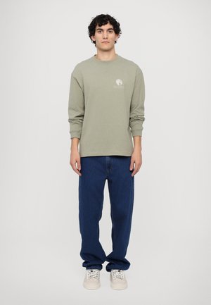 Camicia a maniche lunghe verde oliva con scollo rotondo e dettagli del logo, abbinata a jeans denim blu larghi e sneakers beige.