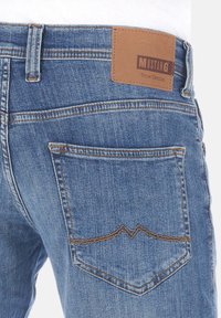 Vaqueros de mezclilla en azul medio con una parche de cuero en la parte trasera etiquetada "MUSTANG True Denim". Presenta costuras en contraste y un diseño clásico de bolsillo.
