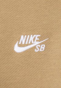 Tanfarget strikket stoff med en brodert hvit "Nike SB"-logo, med en jevn tekstur og et konsekvent mønster over hele stoffet.