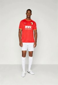 Άνδρας ποδοσφαιριστής στέκεται, φορώντας κόκκινη φανέλα με το λογότυπο WWK, λευκά σορτσάκια, μακριές λευκές κάλτσες και λευκά παπούτσια ποδοσφαίρου σε ουδέτερο φόντο.