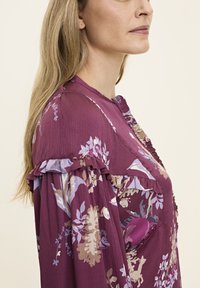Lila Blumenkleid mit Rüschendetails, ausgestattet mit einem geknöpften Ausschnitt und leichtem Stoff. Blumenmuster in sanften Farben überall.