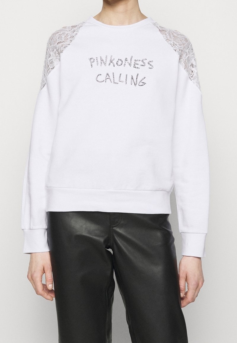 Vit sweatshirt med spetsdetaljer på axlarna, med texten "PINKNESS CALLING" i silverfärg. Mjuk tyg, avslappnad passform, ribbade ärmslut.