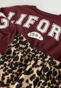 Bordeauxfarbener Hoodie mit weißen „GLIFOR“-Schriftzug und „1982“-Emblem über einem Rock mit Leopardenmuster. Elastischer Bund am Rock.