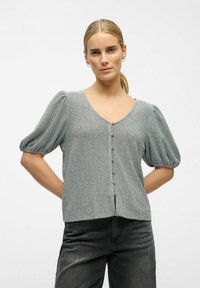 Kvinde med blond hår oppe i en hestehal, iført en grå knapbluse med puffede ærmer og sorte jeans, stående med hænderne bag sin ryg.