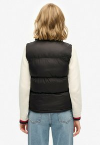 Blonde Person, der eine schwarze gesteppte Weste über einem weißen Pullover mit roten und blauen Bündchen trägt, kombiniert mit hellblauen Jeans und mit dem Rücken zur Kamera steht.