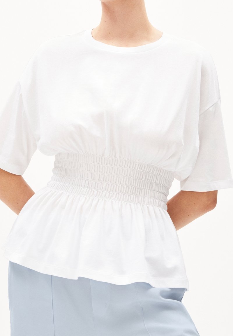 Personne portant un haut blanc à manches courtes avec une taille smockée large et un pantalon bleu clair, les bras positionnés derrière le dos.