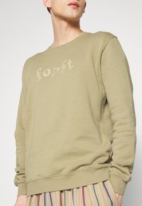 Oliwkowy dres z haftowanym napisem "forêt", okrągłym dekoltem, ribbowymi mankietami i dołem. W zestawie z paskowanymi spodniami w różnych kolorach.