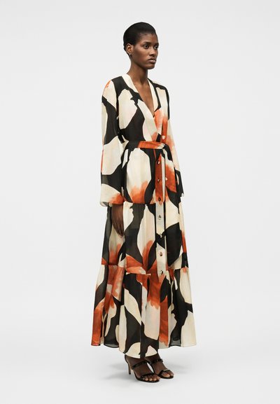 Vrouw staand, gekleed in een lange jurk met ceintuur, voorzien van grote zwarte, witte en oranje abstracte bloemmotieven en zwarte sandalen met hak.