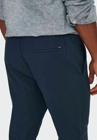 Granatowe spodnie wykonane z gładkiego materiału, z bocznymi kieszeniami, tylną kieszenią z poziomym otworem oraz dyskretną metką marki.