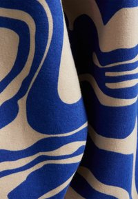 Stickad tyg med vågiga mönster i blått och krämfärg. Texturen verkar mjuk, med en lätt glans som skapar en flytande visuell effekt.