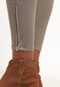 Olivegrønne leggins z zamkiem przy kostce, w połączeniu z brązowymi zamszowymi botkami za kostkę z tasiemką na pięcie.