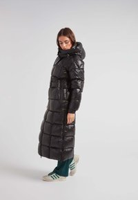 Lange zwarte pufferjas met een glanzende afwerking, voorzien van een capuchon, een rits aan de voorkant en een gequilte stijl, gecombineerd met groene broek en witte sneakers.