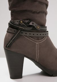 Bota de tobillo marrón de ante sintético con tacón apilado, con correas decorativas de cuero negro y costuras blancas en contraste.