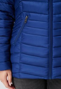Blauwe puffy jas met horizontale stiksels, ritszijvak, staande kraag en een gladde textuur. Sluit met een centrale rits.