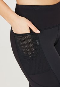 Leggings neri a vita alta con una tasca laterale in rete testurizzata, caratterizzati da un tessuto liscio e dettagli cuciti per una maggiore durata.